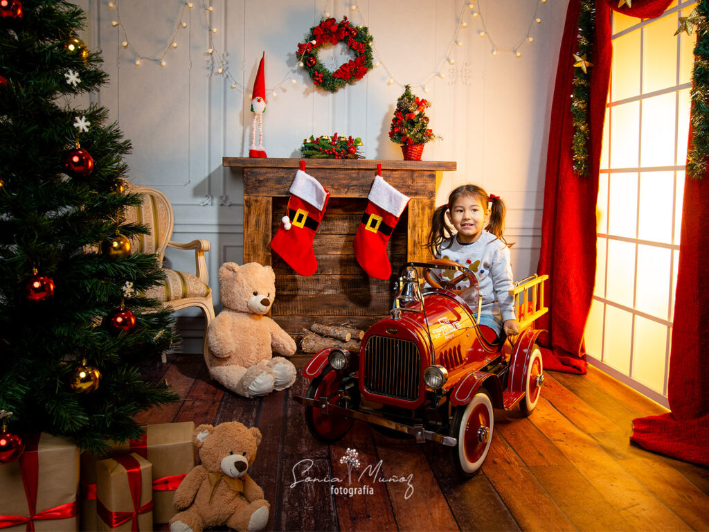 Fotografía Navideña de una niña en un auto que recibió como regalo de navidad junto a un árbol navideño con regalos, el fondo muestra un ambiente acogedor, con decoraciones navideñas, juguetes y una chimenea. © Sonia Muñoz Fotografía, 2020.