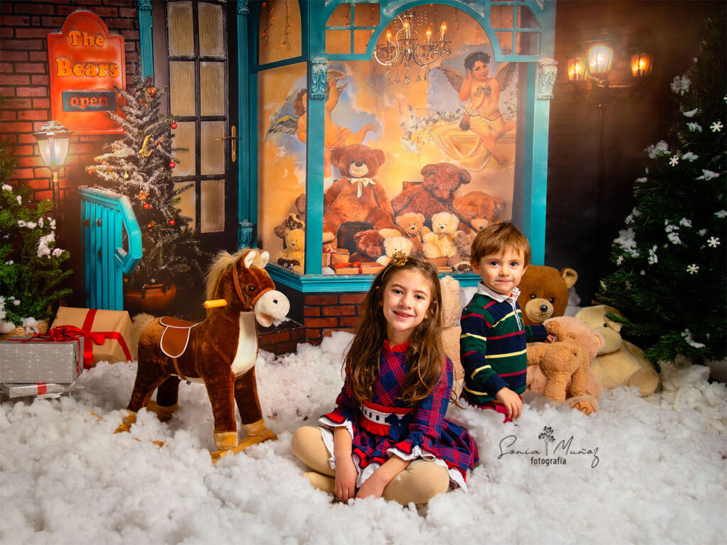 Fotografía de Navidad, de dos niños sobre la nieve, con varios regalos y juguetes de fondo. © Sonia Muñoz Fotografía, 2021.
