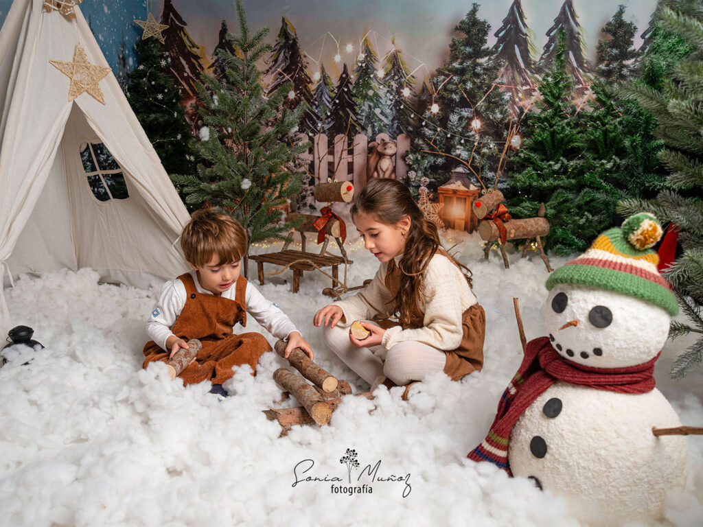 Fotografía Navideña de dos niños jugando con unos troncos rústicos junto a un muñeco de nieve, el fondo muestra una aldea navideña llena de nieve y árboles nevados. © Sonia Muñoz Fotografía, 2022.