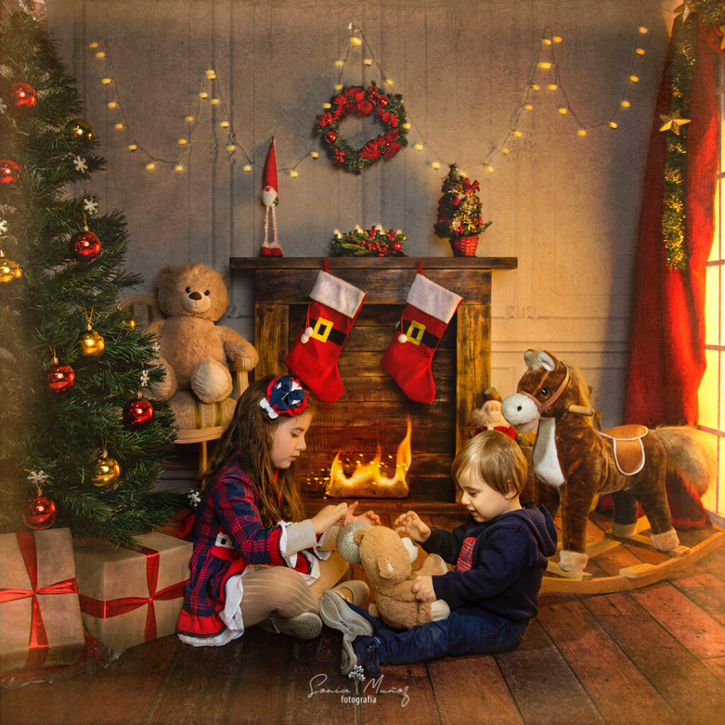 Fotografía Navideña de dos niños jugando junto a un árbol navideño con regalos, el fondo muestra un ambiente acogedor, con decoraciones navideñas, juguetes y una chimenea. © Sonia Muñoz Fotografía, 2020.