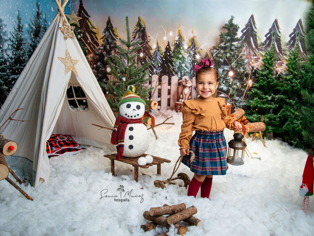Fotografía Navideña de una niña con un trineo, el fondo muestra una aldea navideña llena de nieve y árboles nevados. © Sonia Muñoz Fotografía, 2022.